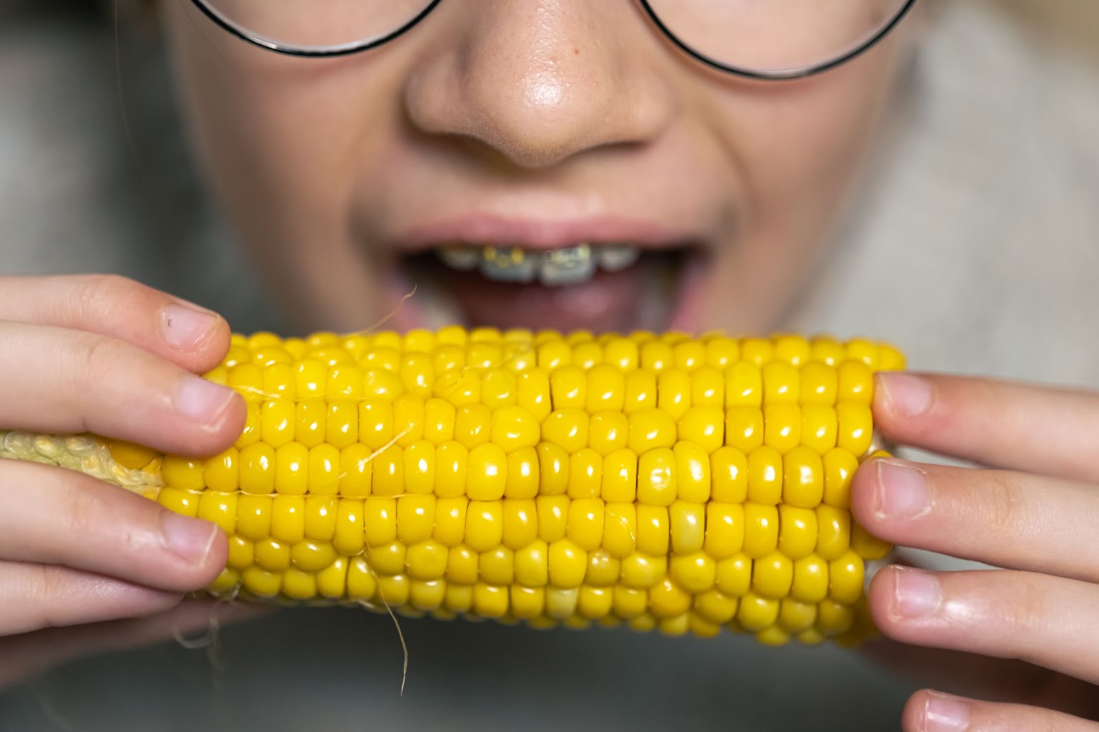 A child with glasses enjoys eating corn on the cob, showcasing a bright smile with straightened teeth from Invisalign aligners.