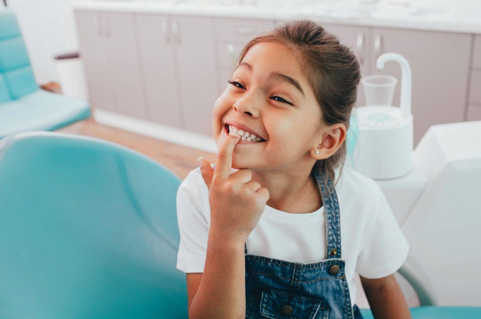 A young girl in a dentist chair rests her finger on her chin, looking calm and curious